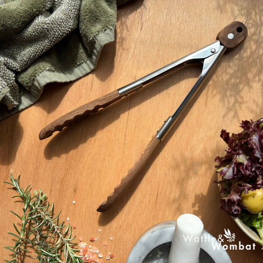 Pair of tongs on a wooden surface with herbs and a mortar and pestle.