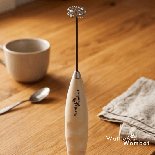 A Wattle & Wombat milk frother with a cream-colored handle and silver metal whisk attachment, standing on a wooden table next to a mug, spoon, and cloth.