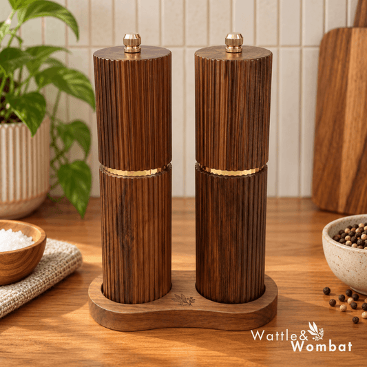 Two ribbed dark wood pepper mills with gold-tone bands and matching wooden bases, shown on a kitchen countertop with bowls of salt and peppercorns and a plant in the background.
