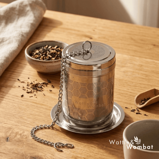 A stainless steel Wattle & Wombat tea infuser with a chain and lid, displayed on a wooden table with tea leaves, a cup, and a cloth napkin.